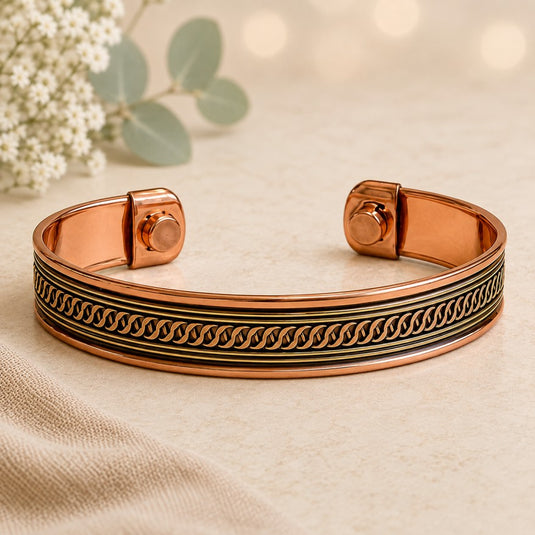 A rose gold cuff bracelet with a black and gold wave pattern, displayed on a beige fabric surface with soft bokeh background and eucalyptus leaves.