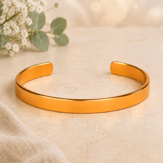 A polished gold-tone open cuff bracelet resting on a light beige textured surface with soft white flowers and green leaves in the background.