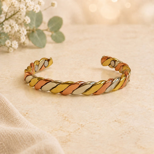 A twisted rope cuff bracelet featuring alternating metallic gold, rose gold, and silver oval links, displayed on a textured beige surface with soft-focus eucalyptus leaves in the background.