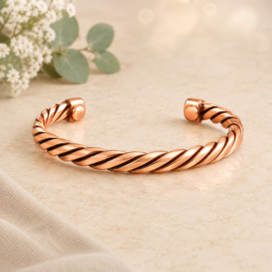 A twisted rope-style cuff bracelet in polished rose gold, displayed on a light beige textured surface with soft-focus white flowers and green leaves in the background.