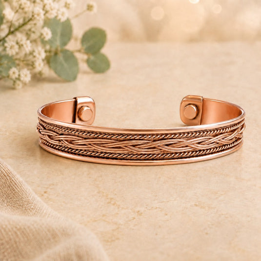 A rose gold cuff bracelet with a detailed twisted rope pattern on the front, displayed on a textured beige surface with soft-focus eucalyptus leaves in the background.