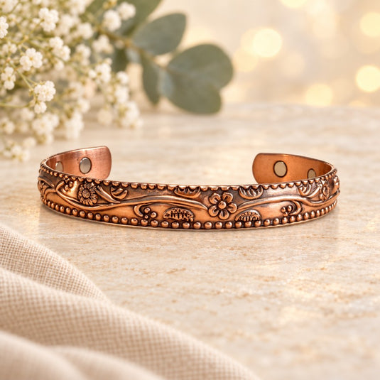 An ornate copper-toned open cuff bracelet with embossed floral and beaded detailing, displayed on a light stone surface with soft-focus flowers and bokeh lights in the background.